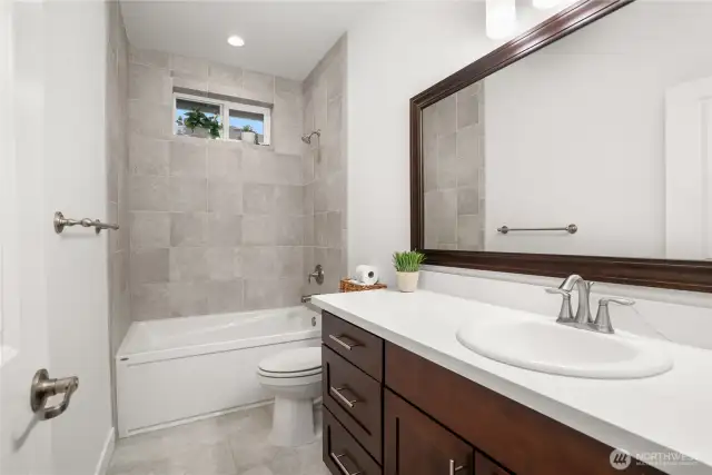 Main level full bathroom featuring a tiled shower-tub combo, quartz countertop vanity, and rich wood cabinetry, conveniently located between the two secondary bedrooms.