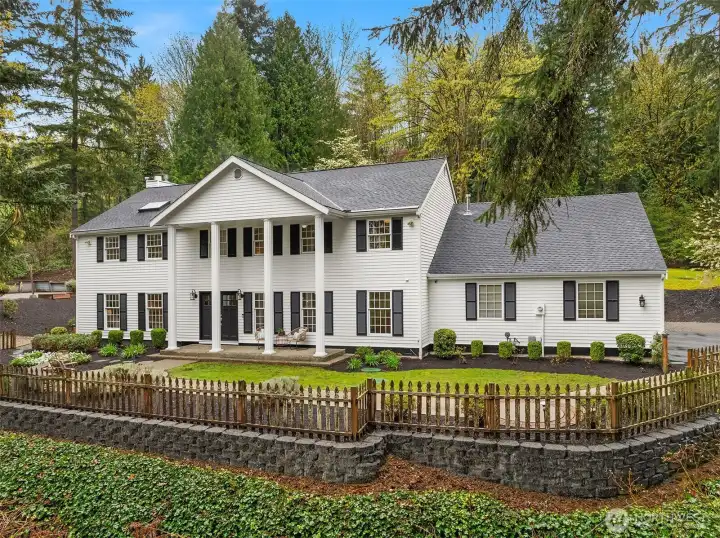 Classic colonial exterior with covered portico, white columns, black shutters, manicured landscaping, and a paver seating area in the foreground