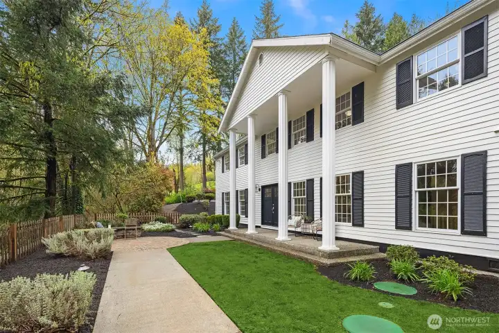 A tree-lined concrete path leads to the covered entry, with manicured hedges and layered plantings framing the colonial facade on all sides