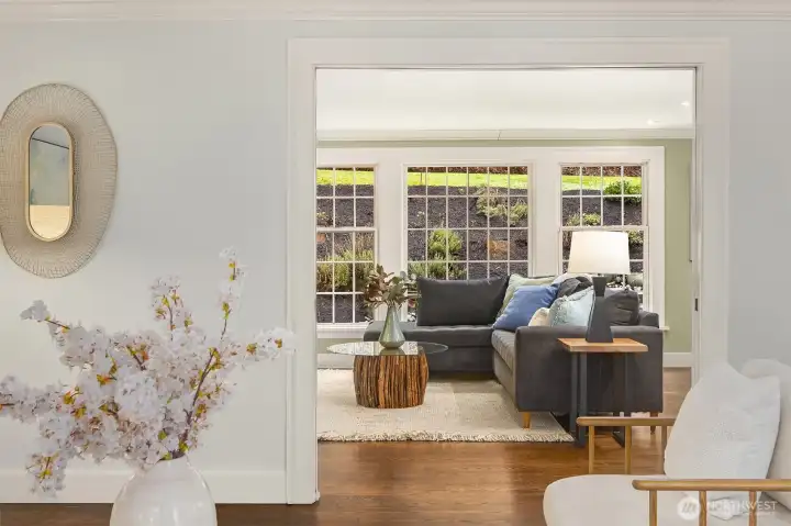 Living room flows into the sun-filled family room beyond — hardwood floors, crown molding, and garden views throughout