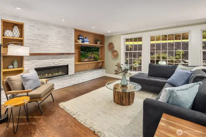 Family room with linear gas fireplace, stacked stone surround, custom built-in shelving, hardwood floors, and large windows overlooking the landscaped grounds