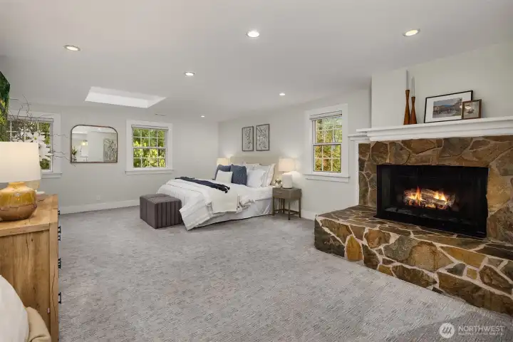 Expansive primary suite with stone fireplace, skylight, recessed lighting, and lush tree-lined views from multiple windows