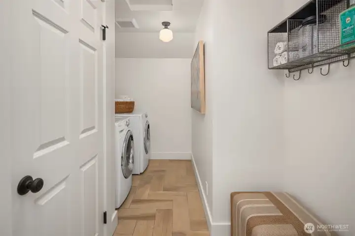 Upper-level laundry room with full-size washer and dryer, herringbone tile floors, wall-mounted storage, and a clean, well-organized layout