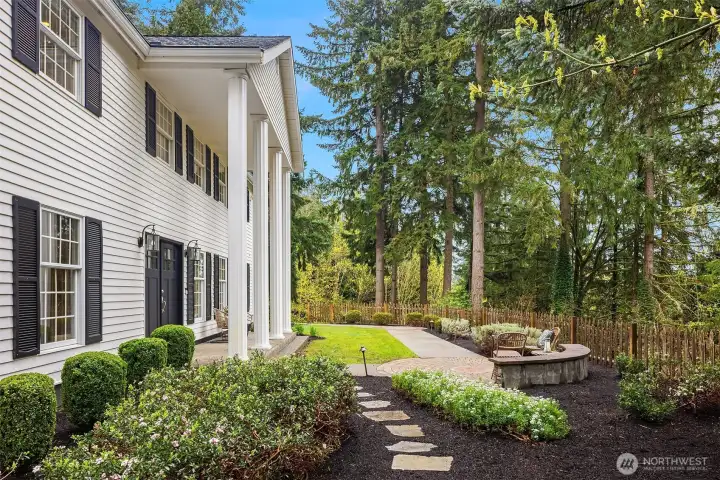 Side exterior view showcasing the colonial architecture, columned entry, mature evergreen privacy, stone path, and seating area
