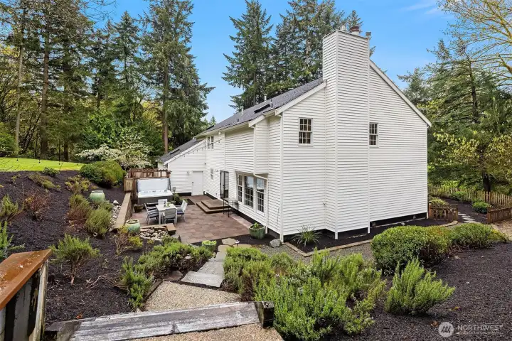 Elevated view of the rear grounds showing the full scope of the property — patio, hot tub, pergola, terraced plantings, and a sweeping lawn framed by towering trees