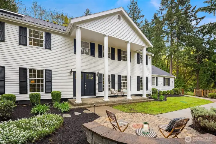 Classic colonial exterior with covered portico, white columns, black shutters, manicured landscaping, and a paver seating area in the foreground