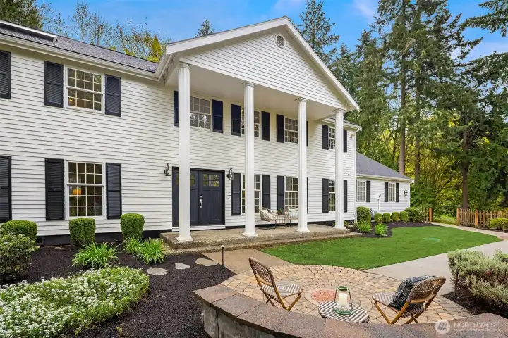 Classic colonial exterior with covered portico, white columns, black shutters, manicured landscaping, and a paver seating area in the foreground