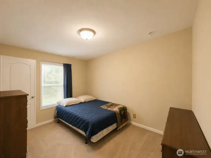 This inviting guest bedroom features soft neutral tones, plush carpeting, and a large window that fills the space with natural light while offering peaceful views of the surrounding property. The efficient layout provides room for bedroom furnishings and personal storage, making it ideal for guests, family members, or flexible everyday living. A warm, welcoming space that complements the home's comfortable country setting.