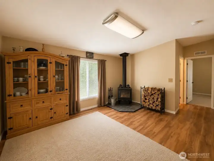 The heart of the home features a cozy freestanding wood stove set on a tiled hearth, creating a warm and welcoming atmosphere during the cooler months. Large windows bring in natural light and offer peaceful views of the surrounding property, while durable flooring and an open layout provide easy flow between the living spaces and bedroom wing. The combination of comfort, functionality, and country charm makes this a perfect gathering space for everyday living and entertaining.
