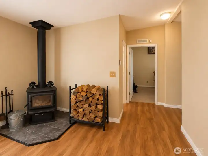 A freestanding wood stove creates a warm and inviting focal point, adding both charm and supplemental heating to the home's open living area. Easy-care flooring, neutral finishes, and a functional layout provide a comfortable transition between the living spaces and bedroom wing. The dedicated wood storage rack and hearth area highlight the home's practical, country-inspired appeal while creating a cozy atmosphere year-round.