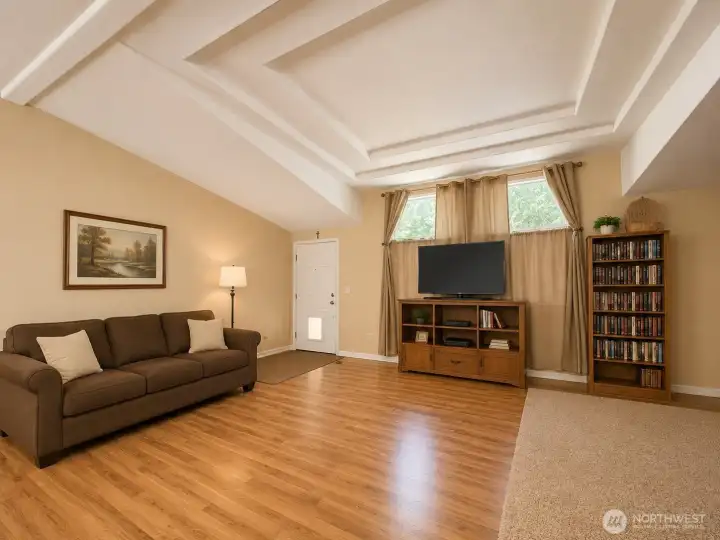 Spacious great room showcasing a striking coffered ceiling, oversized windows, and abundant natural light. The open design creates an inviting gathering space with warm laminate flooring, generous seating areas, and seamless flow to the kitchen and dining room. Architectural details and soaring ceilings enhance the sense of space, making this the perfect centerpiece for relaxing, entertaining, and everyday living.