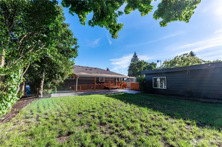 A private, fenced backyard with mature trees, green lawn, and a patio area that connects seamlessly to the deck above.