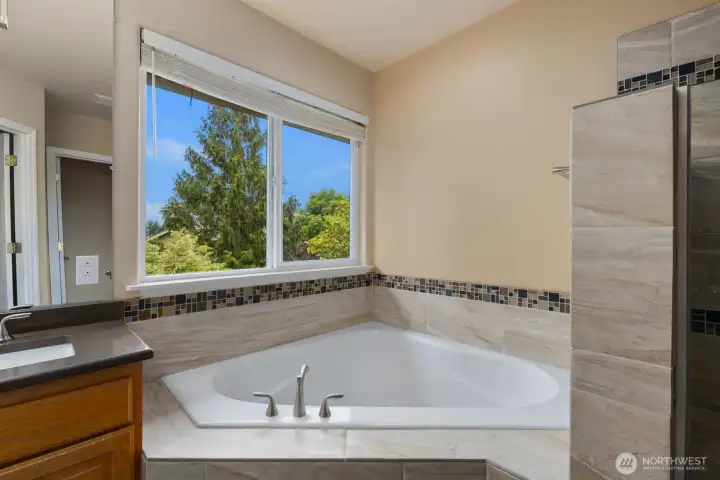 Corner soaking tub positioned beneath a large window, bringing natural light into the primary bathroom. A comfortable space to unwind while enjoying views of the surrounding greenery. Corner soaking tub positioned beneath a large window, bringing natural light into the primary bathroom. A comfortable space to unwind while enjoying views of the surrounding greenery.