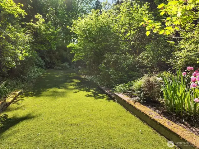 The turf area is bordered by mature trees, blooming flowers, and lush vegetation that changes with the seasons. Plenty of space for new adventures too—perfect for anyone with a green thumb looking to grow their own garden.