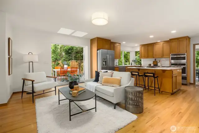 The open flow continues into the kitchen, where hardwood floors and expansive windows create a light-filled space surrounded by lush greenery.
