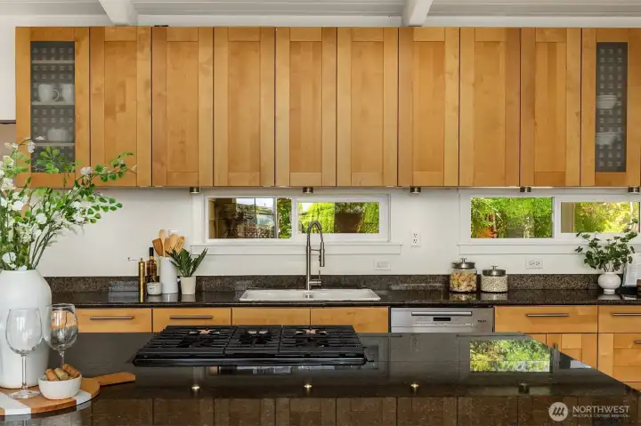 Warm wood cabinetry, expansive counters, and garden-view windows bring a natural warmth to the kitchen, while the oversized island with integrated cooktop creates an inviting space for gathering and everyday living.