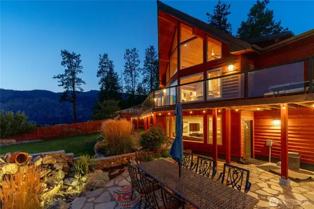 Twilight view of home w/ oversized windows, view decks, outdoor living area w/ pool, hot tub, basketball court, dining and lounge areas, landscaped grounds and water feature.