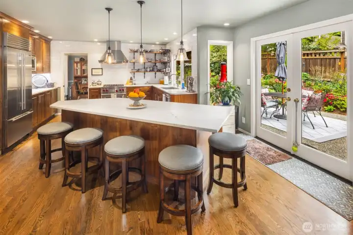 Large rectangular island with a white quartzite countertop and rich wood cabinetry below. 5 round cushioned bar stools line one side, making it a functional space for casual dining or gathering