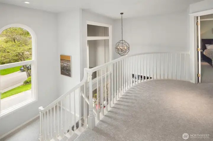 White staircase railing wraps around the opening to the floor below, creating an elegant architectural feature and providing a view down into the entryway. The space is carpeted in a light neutral tone, adding softness and warmth.