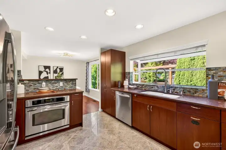The updated kitchen is a chef's dream with custom dark cabinetry, durable quartz counters, stainless appliances, and an artisan glass tile backsplash that brings color and texture to daily prep. A wide garden window above the sink frames lush backyard greenery and the pergola beyond, making dishwashing feel a little less like a chore.