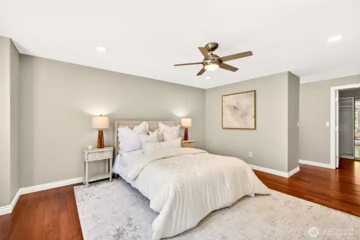 Another angle of the spacious primary suite reveals the generous footprint, gleaming hardwood floors, and direct access to the walk-in closet visible through the open doorway. Calming neutral paint, dimmable wafer LED lighting, and a quiet ceiling fan create the perfect retreat at the end of a long day.