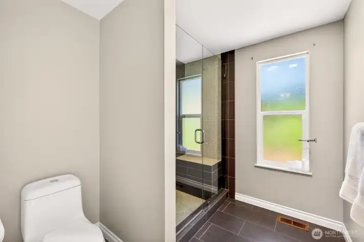 The primary bath continues with a frameless glass walk-in shower enclosure, modern tile work, and a privacy window that floods the space with natural light. Sleek dark tile flooring and clean lines deliver a hotel-inspired atmosphere rare in homes of this era.