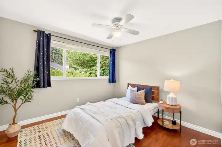 A bright secondary bedroom with rich hardwood floors and a quiet ceiling fan looks out over leafy treetops, making this the kind of cheerful guest room or kid's room everyone wants to wake up in. Wafer LED lighting with day/night toggle adds modern flexibility to a comfortable, well-proportioned space.