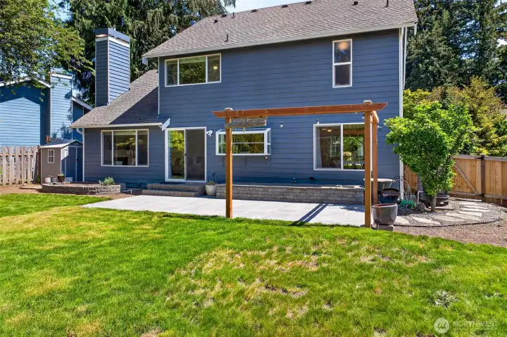 A wider view of the backyard showcases the home's two-story footprint, the wraparound lawn, and the dedicated outdoor storage tucked discreetly along the fence line. With multiple patio zones, mature plantings, and quiet privacy, the outdoor space functions as a true extension of the home's livable square footage.