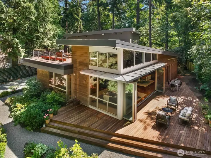 Shed rooflines, walls of glass, and a wraparound ipe deck define the rear elevation. Folding glass doors open the main living space to outdoor dining and lounge areas beneath a mature evergreen canopy.