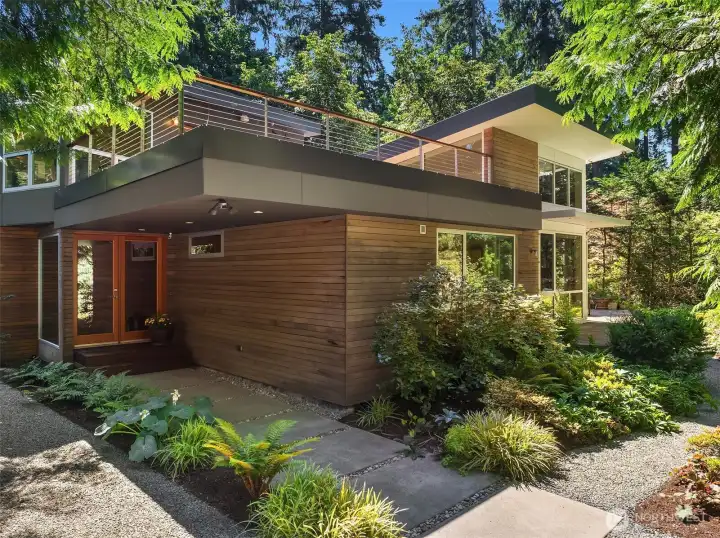Cedar siding and a glass-paneled entry sit beneath the cantilevered roof deck, with a gravel paths winding through established gardens.