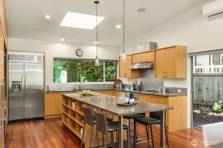 The kitchen centers on a quartz-topped island with seating and open shelving, lit by a skylight and a garden-view window over the sink.