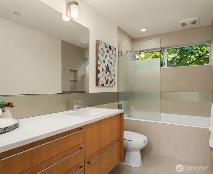 A full bath with floating wood vanity, quartz counter, and tiled tub-shower with frosted glass panel and garden-view window.