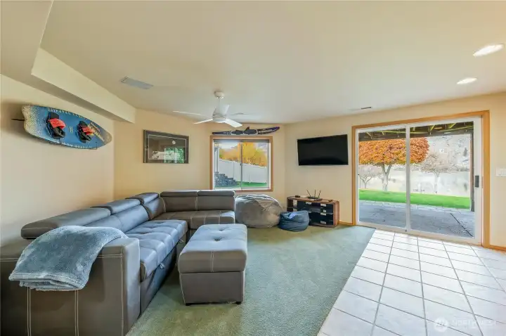 Bright and inviting lower-level rec room with large windows and slider to the patio and lawn. Features plush carpeting, ample seating, and wall-mounted TV — perfect for movie nights and indoor-outdoor living along the riverfront.