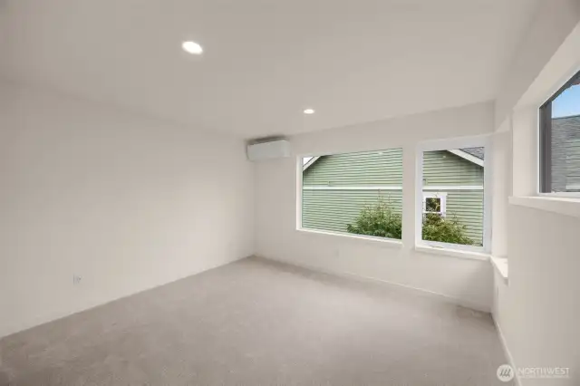 A peaceful bedroom retreat with plush carpet, ductless mini-split, and large windows framing neighborhood greenery.