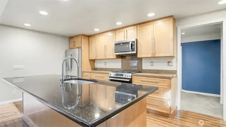 Modern kitchen with natural toned cabinetry. Spacious island features a sink, dishwasher, outlet, and plenty of storage—perfect for meal prep and casual gatherings.