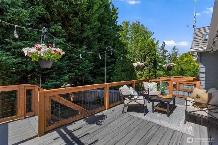 Spacious back deck with composite decking, wood and wire railings, string lights, and hanging flower baskets, a sun-soaked outdoor retreat surrounded by mature evergreens and privacy landscaping.
