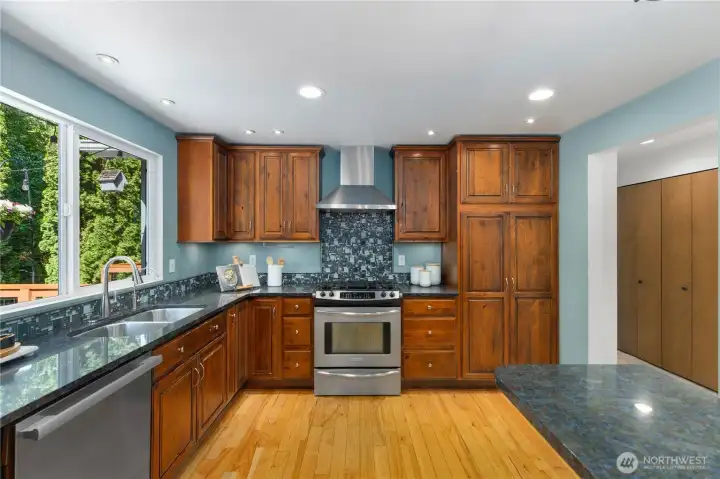 Well-appointed kitchen featuring rich alder cabinetry, dark granite countertops, a mosaic tile backsplash, stainless steel appliances, and a stainless range hood, all set against serene blue-green walls and hardwood floors.