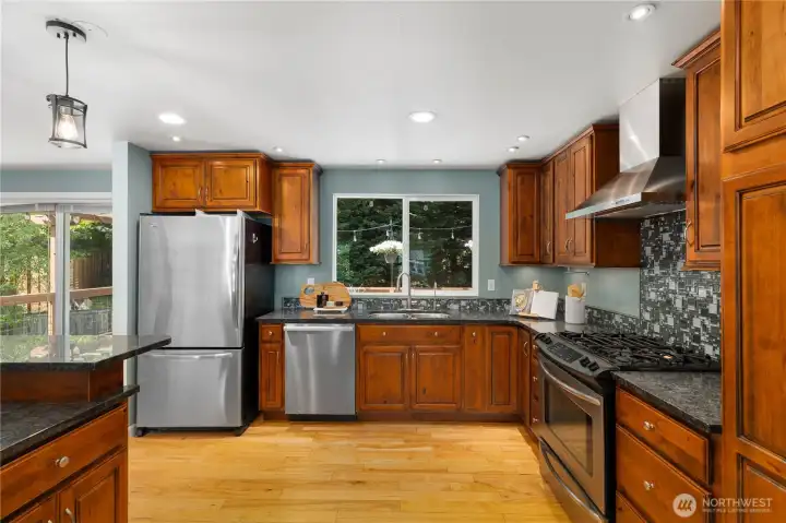 A second angle showcases the full appliance suite including a stainless refrigerator, gas range, and dishwasher, with a garden window above the sink framing lush backyard greenery.