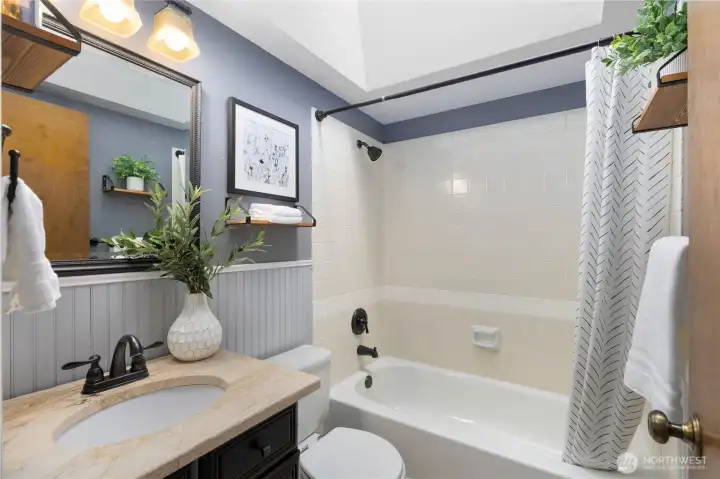 Stylishly updated hall bath featuring wainscoting, a dark vanity with stone-look countertop, oil-rubbed bronze fixtures, tiled tub/shower surround, and a skylight for added brightness.