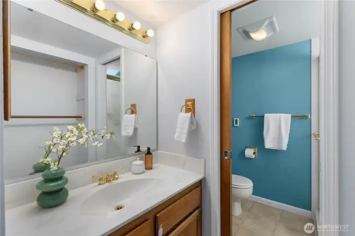Primary 3/4 bathroom with a wide single-sink vanity, large wall mirror, gold fixtures, and a private water closet with teal accent wall, bright and neatly presented.