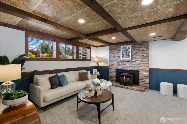 A character-rich lower-level family room featuring exposed wood beam ceilings, bold navy wainscoting, a brick fireplace, and garden-level windows, a warm and inviting bonus space with great flex potential.
