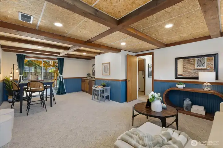 A wide-angle view of the spacious lower level reveals a generous multi-use layout with room for living, dining, and play, plus a wet bar area, sliding door to the backyard, and exposed beam ceiling throughout.