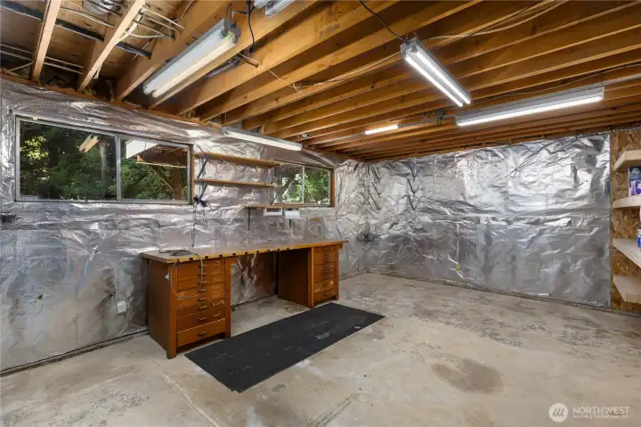 A large unfinished workshop area behind the garage with concrete floors, reflective insulation, built-in wood workbench, open shelving, and bright fluorescent lighting, ideal for hobbies, projects, or additional storage.