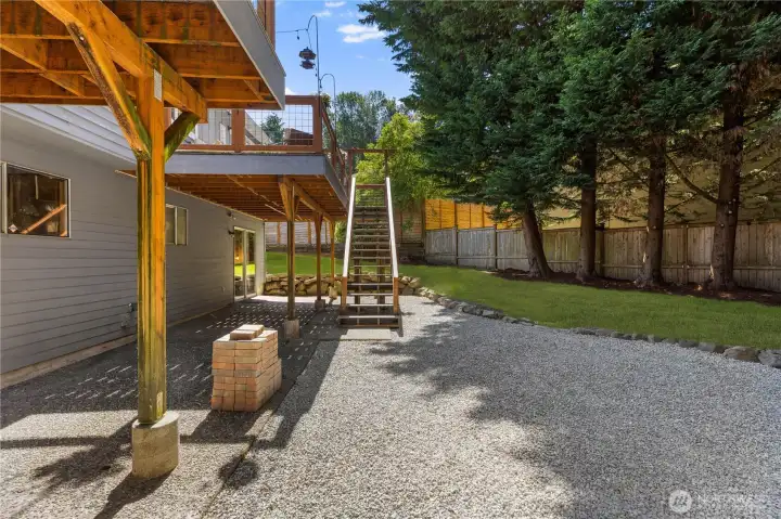 The lower level of the backyard features a gravel patio beneath the elevated deck, with staircase access to the upper deck, lower-level sliding door entry, and a fully fenced yard, a functional outdoor space with flexible use potential.