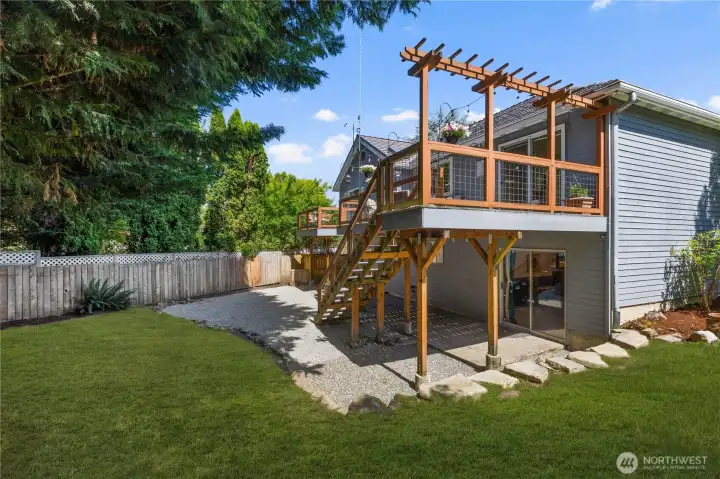 A view from the yard captures the full scale of the elevated deck with pergola overhead, wire-panel railings, staircase to grade, and a lower patio below, showcasing the home’s impressive multi-level outdoor living setup against a lush, fenced backdrop.