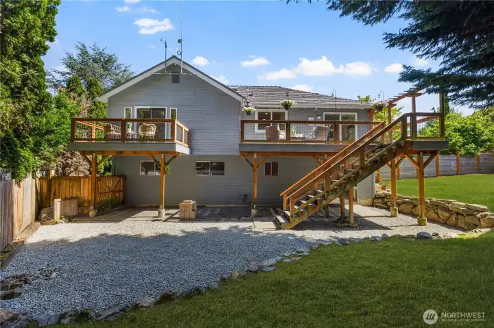 Full rear view of the home highlighting the expansive elevated deck with dual seating areas, staircase access, lower gravel patio, and generous lawn, a backyard designed for both entertaining and everyday enjoyment.