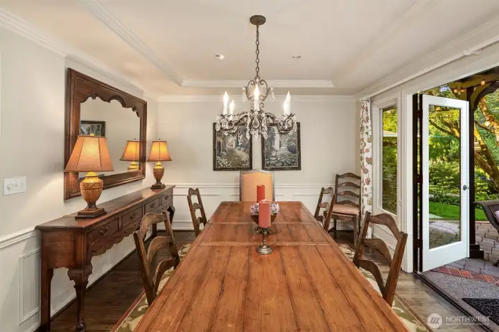 Dining room with tray ceiling and French doors leading out to a patio surrounded by lush landscaping.