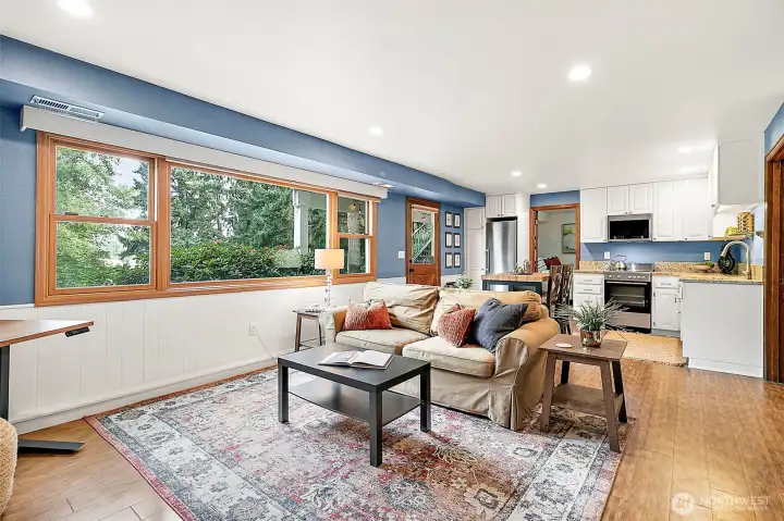 Bright and inviting open-concept living space with a full kitchen, dining, and cozy seating area, all framed by expansive windows that bring in natural light and capture views of the surrounding greenery.