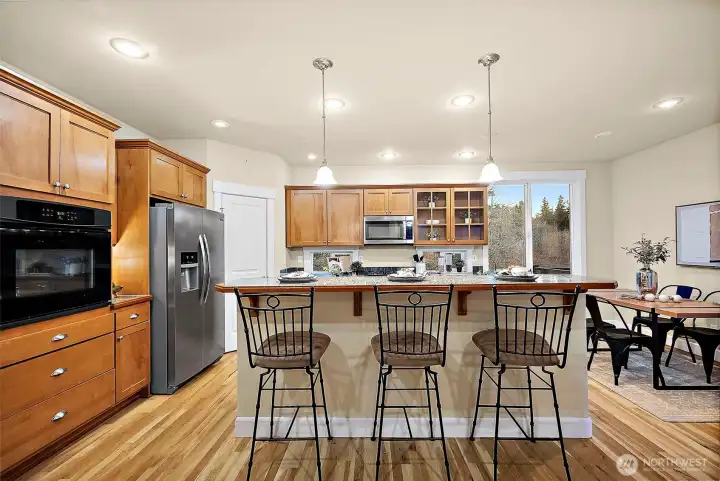Island with bar seating, in-wall oven, side-by-side refrigerator, wood cabinetry and hardwood floors
