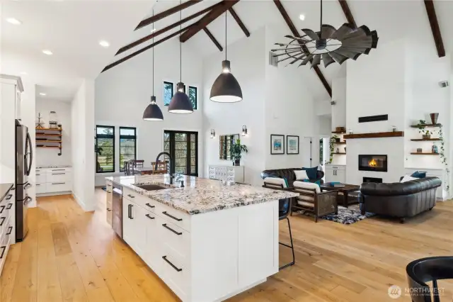 Gourmet kitchen with huge island will accommodate enough space for 4-6 barstools. Tasteful pendant lighting above the island and alpine white granite graces the countertops. This photo shows the soaring, wood beamed ceilings, custom fan, living, dining and kitchen as well as a peek into the large pantry.